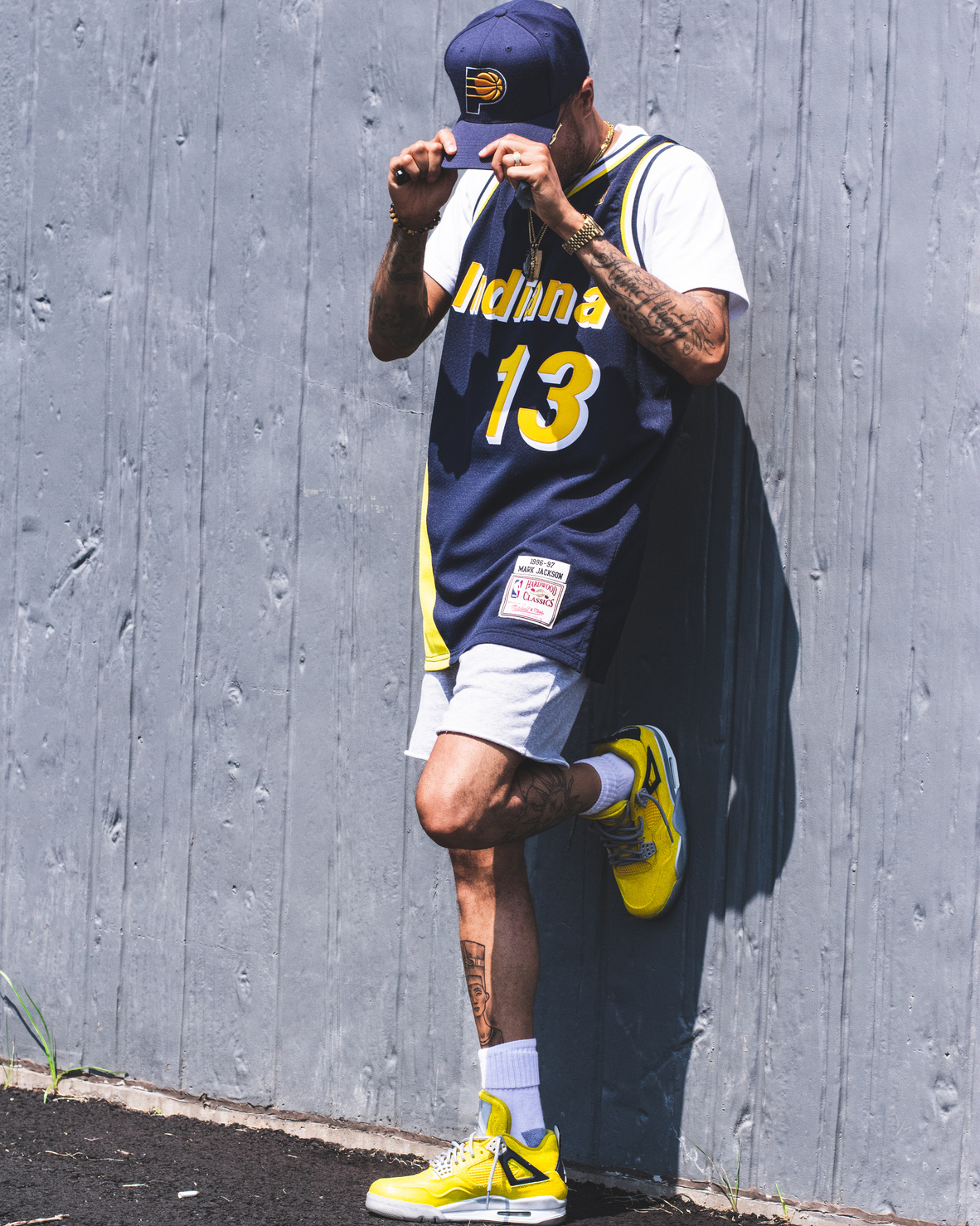Photo of Man in Blue and Yellow Indiana Basketball Jersey and Gray Shorts Leaning on Gray Wall While Looking Down